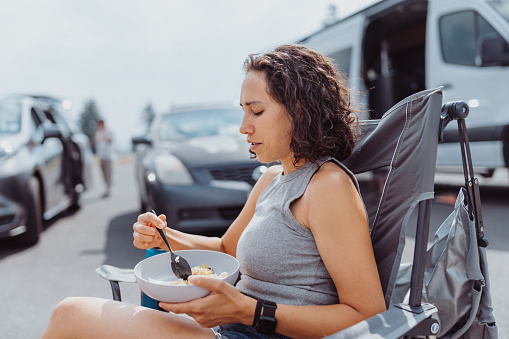Profile view of an Eurasian woman sitting in a camping chair and eating lunch outside her camper van while on a road trip with her family.