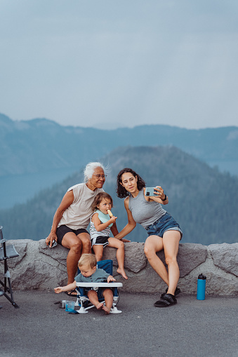 An Eurasian woman uses her smart phone to take a selfie photograph with her toddler daughter and her vibrant senior adult mother who is of Hawaiian descent. The multi-generation family is relaxing at a scenic viewpoint during a camping road trip and Crater Lake is visible in the background.