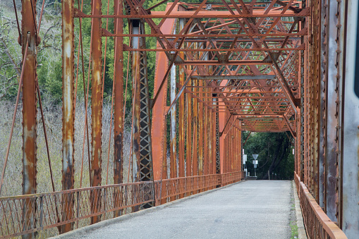 Looking down the roadway on an old red metal bridge. The bridge girders are on the top and sides. The road is paved. Trees are at the end of the bridge.