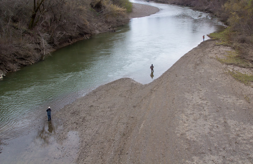 3 fishermen are fishing in a river. One is in the water and other two on on the sandy beach. Trees are across the river. The photo is looking down on them.