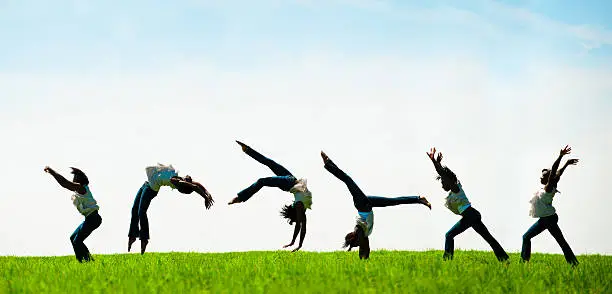 Talented African American woman performs back flip through a scenic grassy field with blue sky. She is a gymnast and demonstrates perfect form with pointed hands a toes.