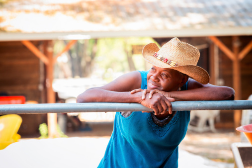 Mature Black Woman Resting on Ranch Fence and looking left with a smile. She is wearing a straw cowboy hat and a blue top.
