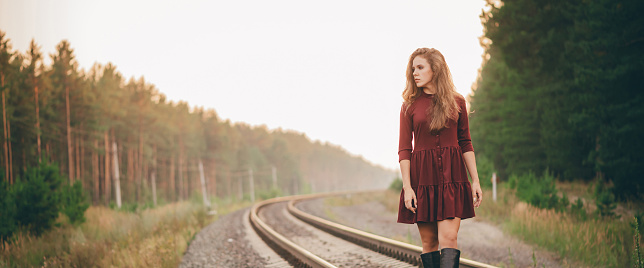 Beautiful dreamy girl with curly natural hair enjoy nature in forest on railway. Dreamer lady in burgundy dress walk on railroad. Inspired girl balancing on train rail at dawn. Sun in hair in autumn.