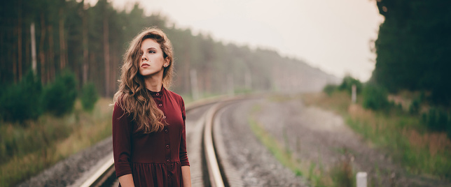 Beautiful girl with curly natural hair enjoy nature in forest on railway. Dreamer lady in burgundy dress walk on railroad. Female portrait of inspired girl on rails at dawn. Sun in hair in autumn.
