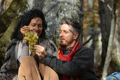 A beautiful Latina woman and a white man, happy, in love and smiling, have fun on an autumn trip in the forest and eat grapes.\nThe man has a red scarf.