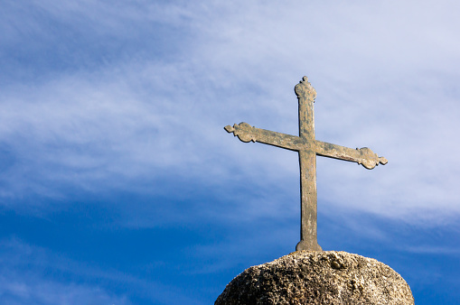 Metallic catholic cross on rock pedestal, with a blue sky with clouds in the background. Copy space.