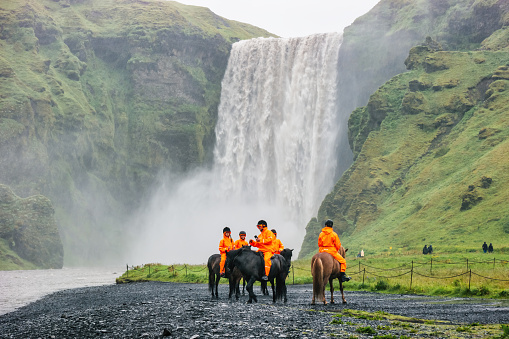 People take part in a horse-riding tour at Skogafoss waterfall in Iceland on a rainy day.