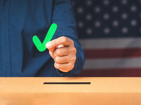 Hand holding a green check mark symbol overhead the voting box at place election with an American flag background. Freedom democracy concept. Campaign to exercise the right to vote