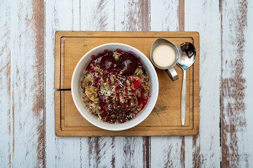 Young adult girl making oatmeal for breakfast, with fresh organic fruit, nuts and grains, at a wooden table in a modern white kitchen, representing a healthy lifestyle, an image with a copy space