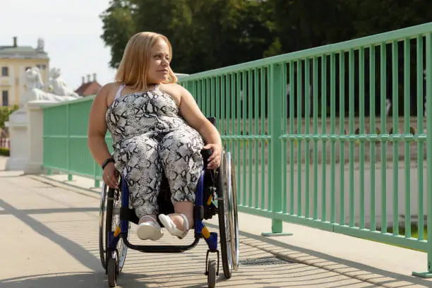 Beautiful Young Woman With Short Height Walks Over Bridge In Wheelchair In City at Summer Day. Female Adult With Disability. Copy Space For Text. Horizontal Plane.