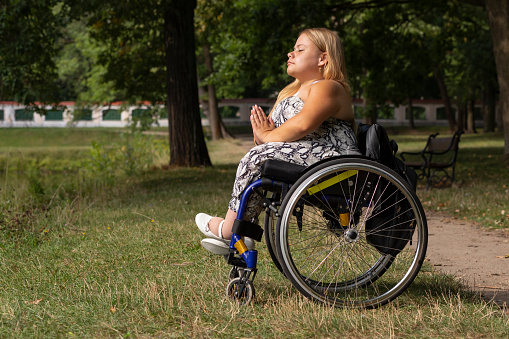 Young Woman With Short Height On Wheelchair Meditates, Relaxing in Green Park At Summer Day. Full Length Female Adult With Disability Enjoys Time In Nature. Horizontal Plane. High quality photo