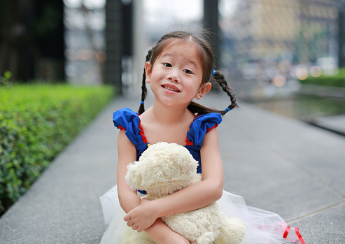 Happy little Asian girl dressed with a fantasy costume sitting in the garden with hugging teddy bear.