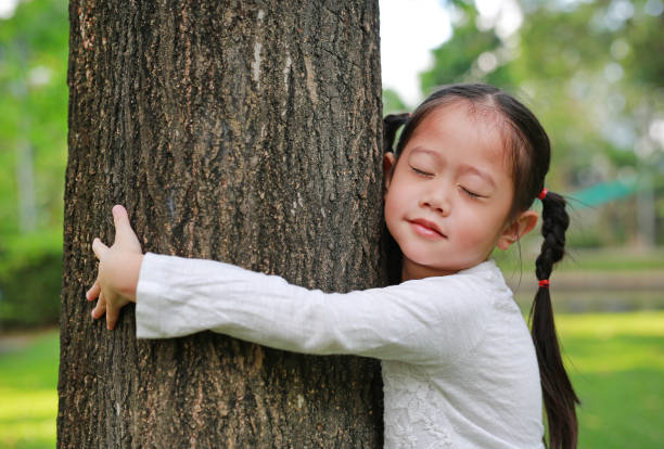 primer plano de una niña sonriente abrazando un árbol con los ojos cerrados en el jardín. - enfold fotografías e imágenes de stock