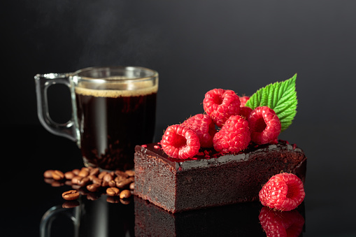 Chocolate cake with fresh raspberries and black coffee on a black reflective background.