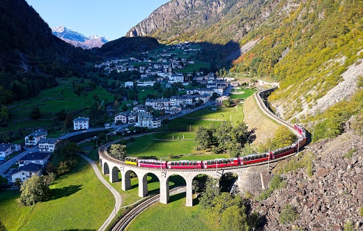 Aerial view of a Bernina Express train crossing the famous Brusio spiral viaduct of Rhaetian Railway over a green grassy meadow with fall colors on the rocky mountainside, in Graubunden, Switzerland