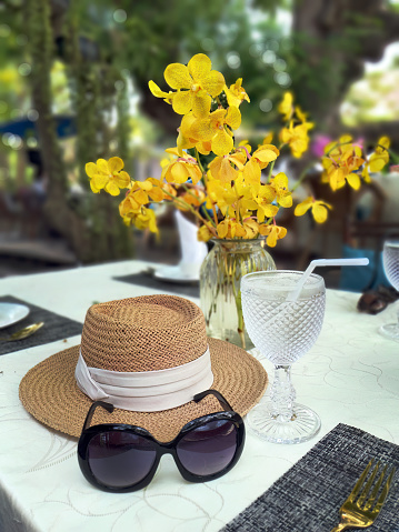 A sunglasses and woven straw sun hat decorated with beige color ribbon on the white color table cloth, lunch table with crystal stemmed glass and a vase of yellow orchids flower, luxury concept