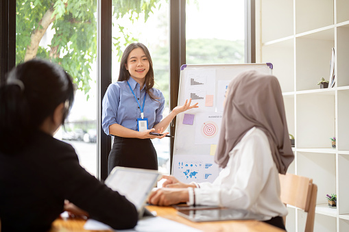 A pretty and charming young Asian businesswoman or female financial assistant is presenting her summarized financial data on the board to her team in the meeting.