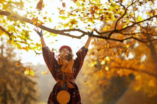 Hello autumn. happy elegant middle aged woman in red hat with scarf, gloves and bag throwing autumn leafs in the city park.