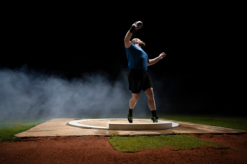Male athlete throwing shot put ball while practising on track at night in arena.
