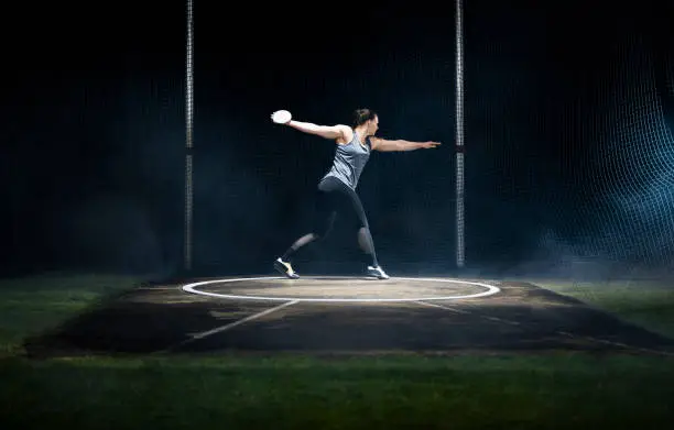 Female athlete turning with discus on track during practice at night in arena.
