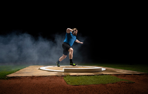 Male athlete holding shot put ball while practising on track at night in arena.