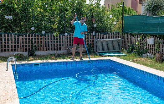 Mature man in flip flops cleaning the swimming pool with a net. Man working as a cleaner of the swimming pool, he standing with special equipment for cleaning at poolside and working