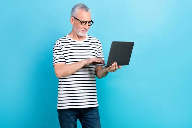 Photo of Photo portrait of funny old male showing laptop apple macbook samsung dressed stylish striped garment isolated on blue color background