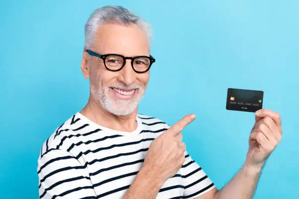 Photo portrait of funny old male credit card finger point empty space dressed stylish striped garment isolated on blue color background.