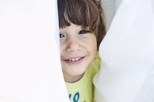 A close up shot of a cute and playful white Caucasian kid smiling and poking his head between white curtains