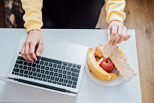 Freelancer young woman eating healthy food when working from home. Woman eating Healthy Grain Snacks and fruits while working with laptop at home office.