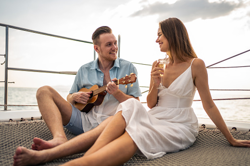 Caucasian romantic couple singing and play ukulele during yachting. Attractive young man and woman hanging out celebrating anniversary honeymoon trip while catamaran boat sailing during summer sunset.