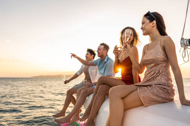 un grupo de amigos diversos beben champán mientras tienen una fiesta en yate. atractivos hombres y mujeres jóvenes pasando el rato, celebrando el viaje de vacaciones mientras navegan en catamarán durante el atardecer de verano. - drinking little girls women wine fotografías e imágenes de stock