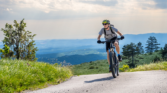 Man riding electric bicycle on road against mountain range and cloudy sky.