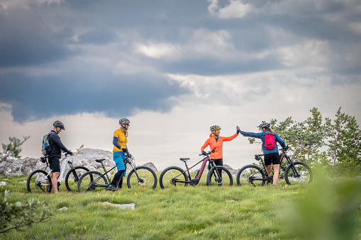 Mature men watching mature women giving high-five to each other while standing with mountain bikes on grassy hill.