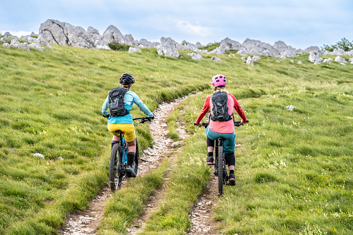 Rear view of female friends riding electric bicycles on grassy mountain trail.