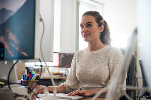 Close-up of a young female office worker sitting at her desk using a computer to complete her work in the office in Newcastle, England. She is wearing smart casual work clothing.