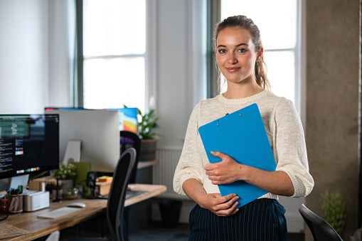 Close waist-up of a young female office worker standing by her desk, she is holding a clipboard in the office in Newcastle, England. She is wearing smart casual work clothing while looking at the camera.