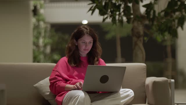 Young woman working in laptop sitting on couch at night, hotel lobby