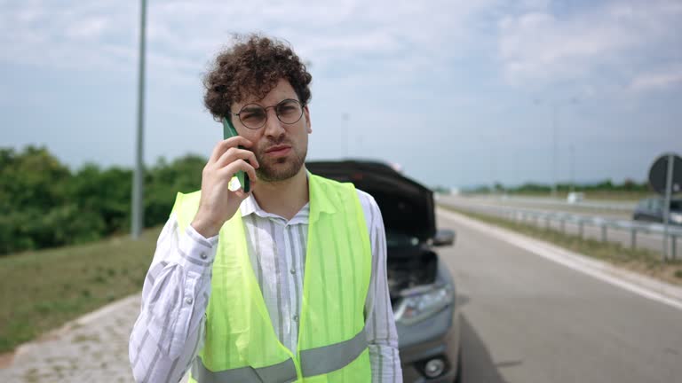 Young handsome man with reflective west calling help for his broken vehicle on highway