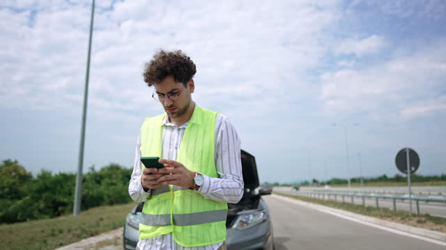 Young man with reflective west text messaging for help for his broken vehicle on highway