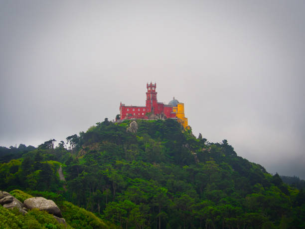 Moors Castle, Sintra, Portugal - Jul 04 2019 : A view of the deep red and bright yellow National Palace of Pena as seen from the heavily fortified Moors Castle on an overcast afternoon.