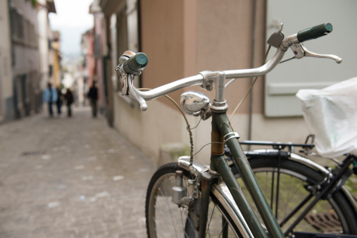 This is a horizontal color photograph of a vintage bike parked alongside a cobblestone street in Zurich, Switzerland. Depth of field is shallow. Focus is on the bell attached to the left handle. Out of focus in the distance people walk down the street. 