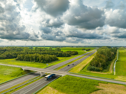 Traffic driving over a highway in a rural landscape with a clouds above in Flevoland, Netherlands. A bridge crosses the highway with traffic driving underneath. The A6 highway is running next to the IJsselmeer lake.