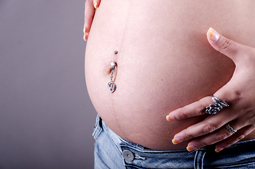 Horizontal studio shot on gray of pregnant mixed race woman's belly with peircing and hands.