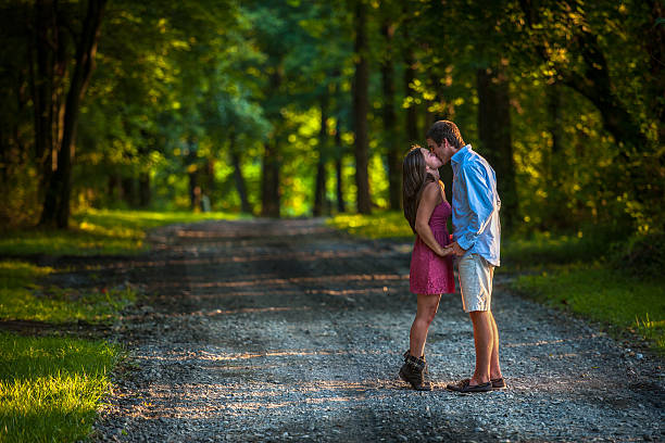 Charming young couple hold hands and kiss as they walk down a gravel road through a wooded setting.