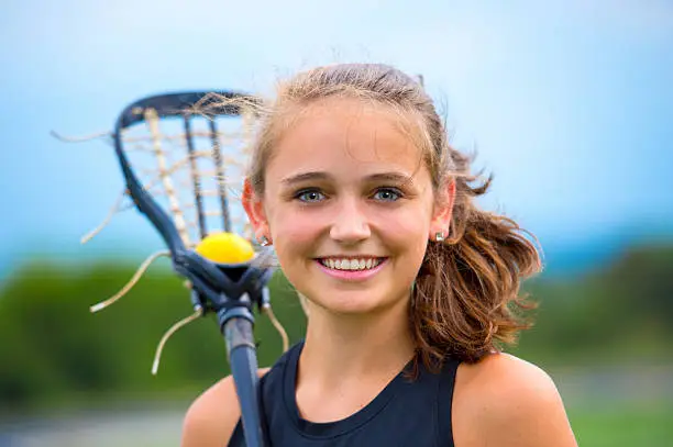 Girls Lacrosse player portrait outside during game. She smiles broadly and makes eye contact with the viewer. She is holding a lacross stick with ball in pocket over her shoulder.