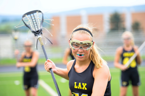 Team captain stares down the viewer with teammates defocused in the background. She is wearing full protective gear including eyes and nose guard and mouth guard. Holding a lacross stick.