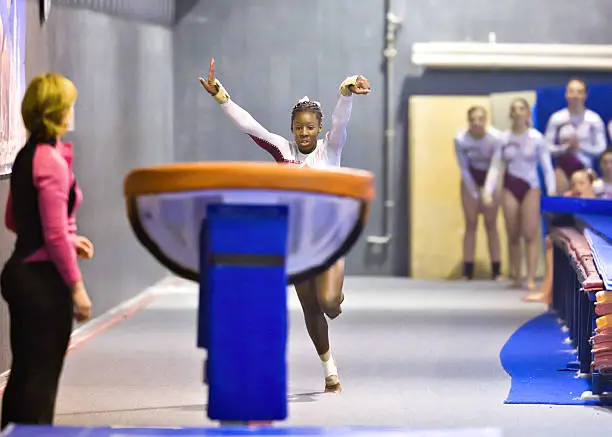 Talented African American gymnast runs towards the vault, with her arms raised as she is about to dive foward to hit the spring board. Her teammates look on in the background and her coach stands close by to spot.