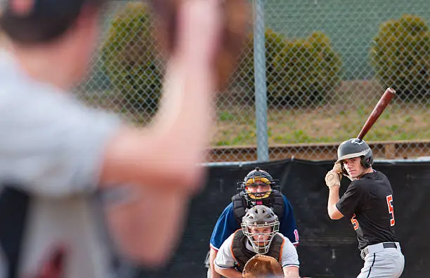 Tight shot framing the pitcher, catcher, umpire, and batter during a baseball game. Shallow selective focus on the batter. Much action in the photo. All eyes concentrate on the pitcher who is mid way through his pitching motion.