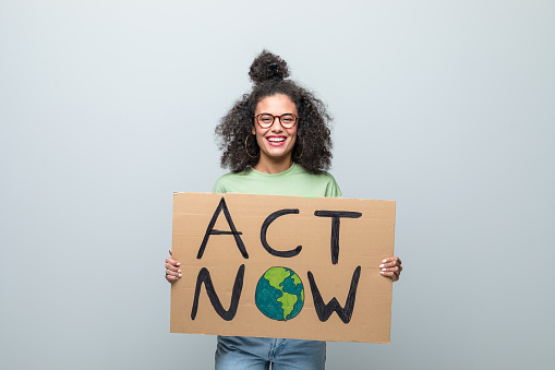 Happy young woman wearing green t-shirt and eyeglasses holding „Act now” poster and smiling at camera. Studio shot against grey background.